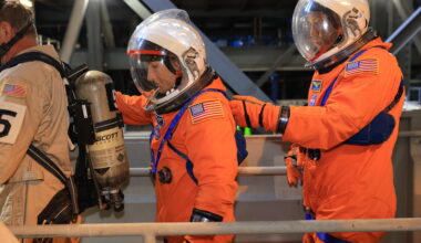 NASA astronauts Christina Koch, a white woman, and Victor Glover, an African-American man, walk to the left during a night-run launch day demonstration. The crew access arm they're walking on has railings, one of which is in the foreground. The astronauts are wearing orange suits with patches and straps on them, as well as white helmets with clear visors. There is a person walking in front of them. This person is wearing khaki clothing and is carrying a gas tank on their back. The gas tank has straps like a backpack.