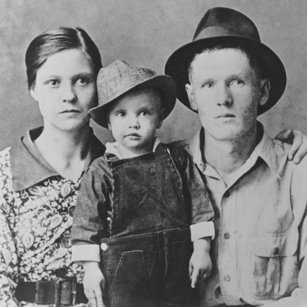 a black and white photo of a man and woman posing with a child, who wears overalls and a fedora