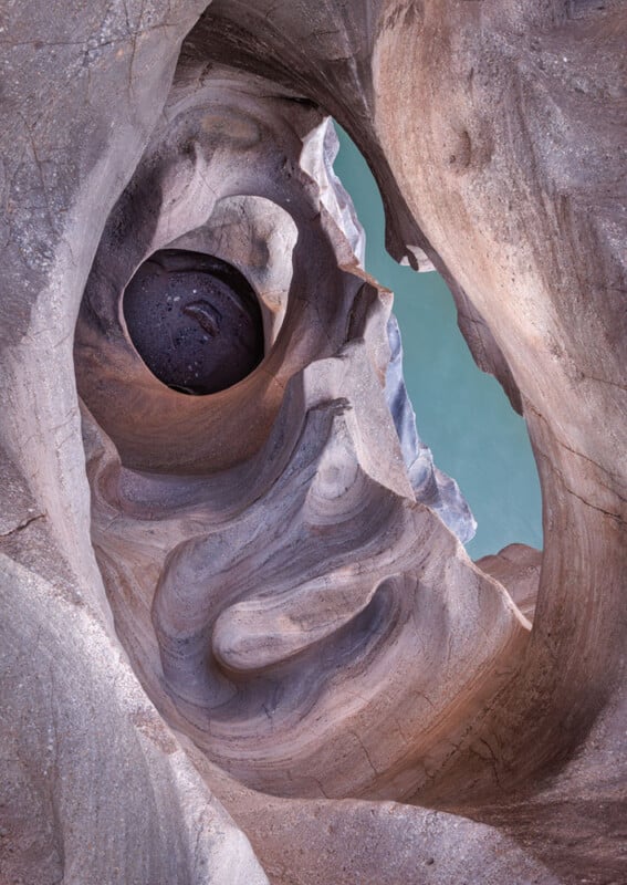 Swirling, smooth sandstone rock formations frame a circular hole through which pale blue-green water is visible below, creating an abstract, organic pattern with flowing lines and curves.