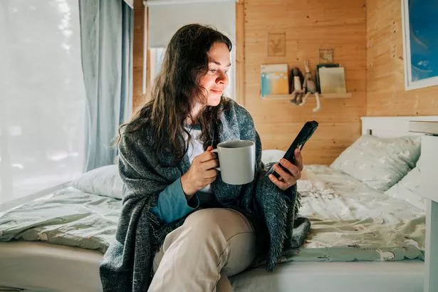 Woman sitting on bed, wrapped in blanket, drinking tea and browsing smartphone in cozy wooden room