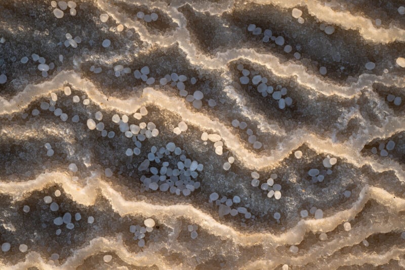 Aerial view of wavy salt formations with scattered round salt crystals on a textured, light brown surface, illuminated by warm sunlight.