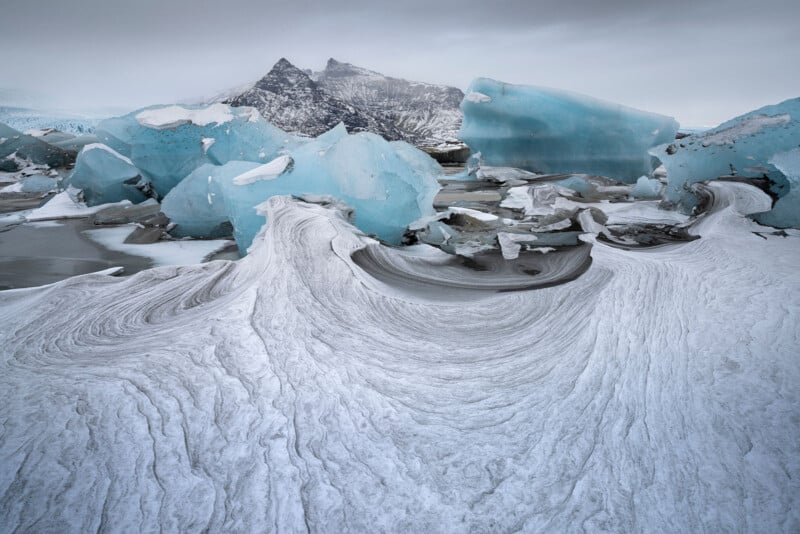 Large, blue icebergs and swirling snow patterns cover a frozen landscape, with rugged mountains rising in the background beneath a cloudy gray sky.