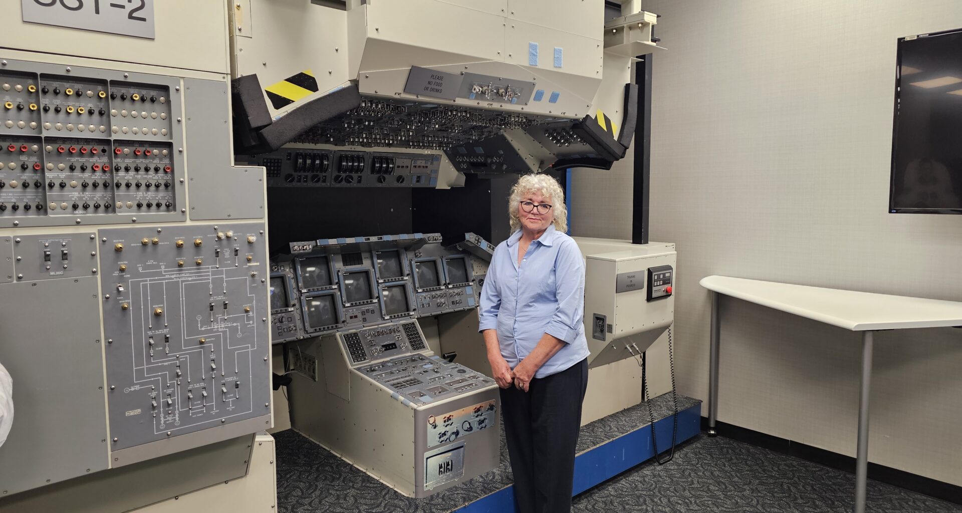 A woman wearing business casual clothing stands in front of an old space shuttle cockpit mockup.