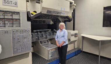 A woman wearing business casual clothing stands in front of an old space shuttle cockpit mockup.