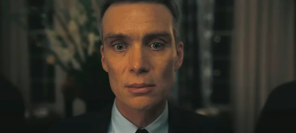 A person in formal attire looks directly at the camera with an intense expression, set against a blurred indoor background