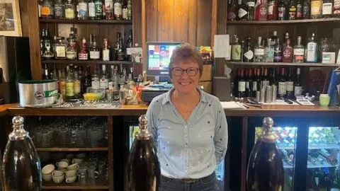 A woman in a light blue shirt standing behind a bar in a pub.