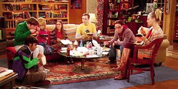 Group of friends sit around a living room table, eating takeout and engaged in conversation