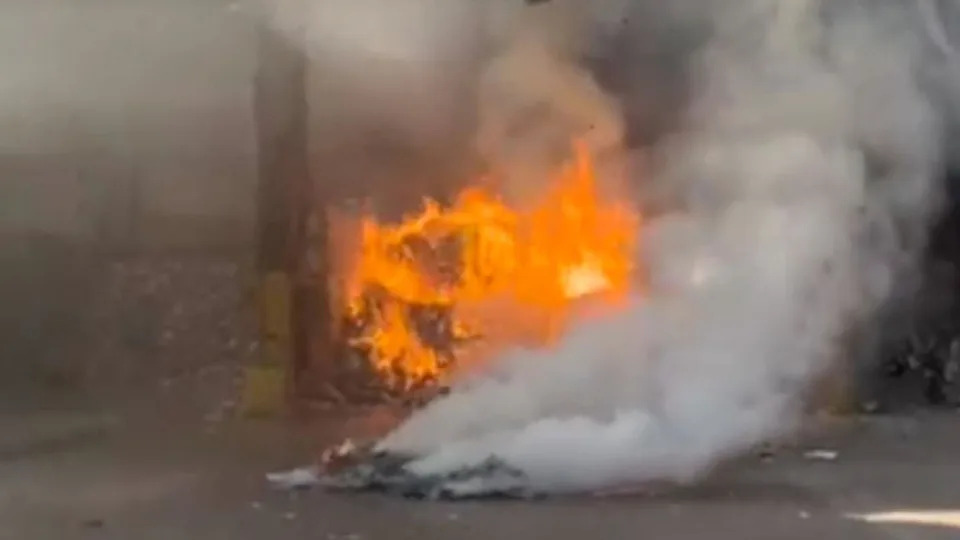 A fire rages in a recycling facility. Plumes of smoke rise in the foreground, in front of a pile of material burning with bright orange flames.
