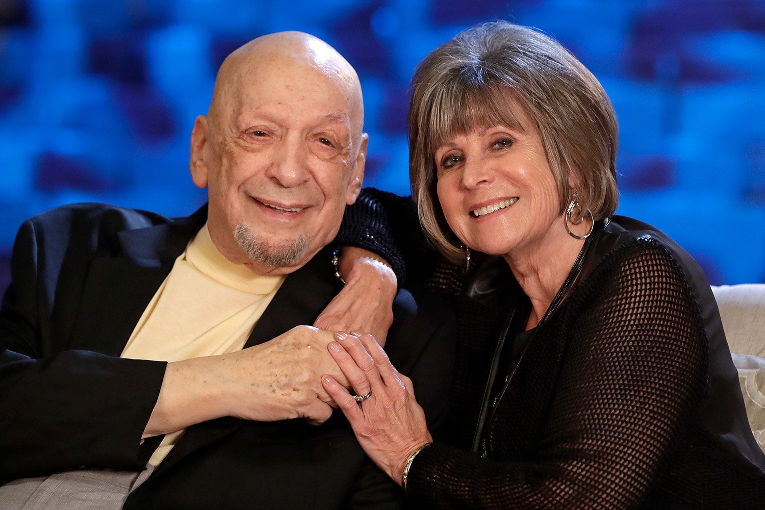 An older man and woman are seated and lean in to put their heads together. 