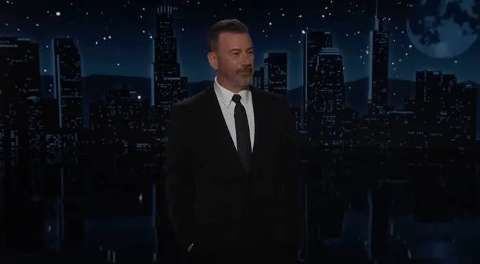 A person in a suit and tie stands on a nighttime cityscape set, likely during a talk show or news segment