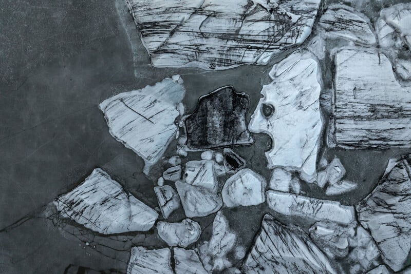 Aerial view of cracked, irregular pieces of ice on a dark, frozen surface, with various shades of white, gray, and black creating abstract patterns across the scene.