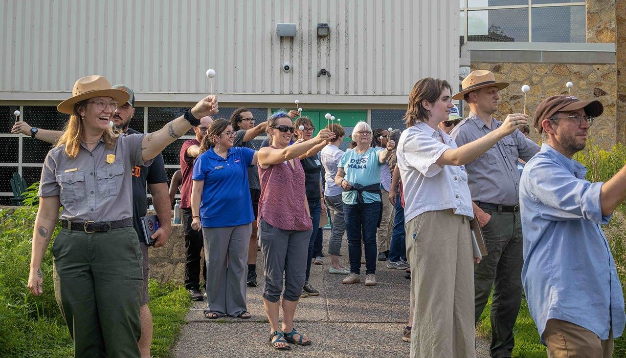 A group of people stand outside holding white Styrofoam balls on a stick with arms outstretched to model moon phases.