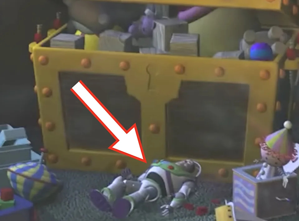 Buzz Lightyear is lying on the floor in front of an open toy chest, surrounded by various toys in a playroom setting