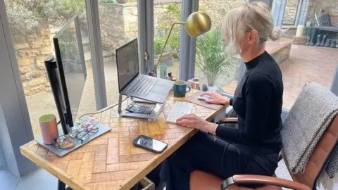 A woman sitting at a desk in a home office. She is wearing a black turtleneck and blonde hair.

On the desk are several pairs of glasses, a monitor, a calculator, a laptop on a stand, a phone and a mug.