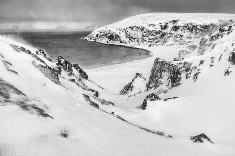 Snow-covered cliffs surround a bay with a sandy shore. Snowfall blurs the sky and the distant hills, creating a cold, windswept, and dramatic winter landscape in black and white.