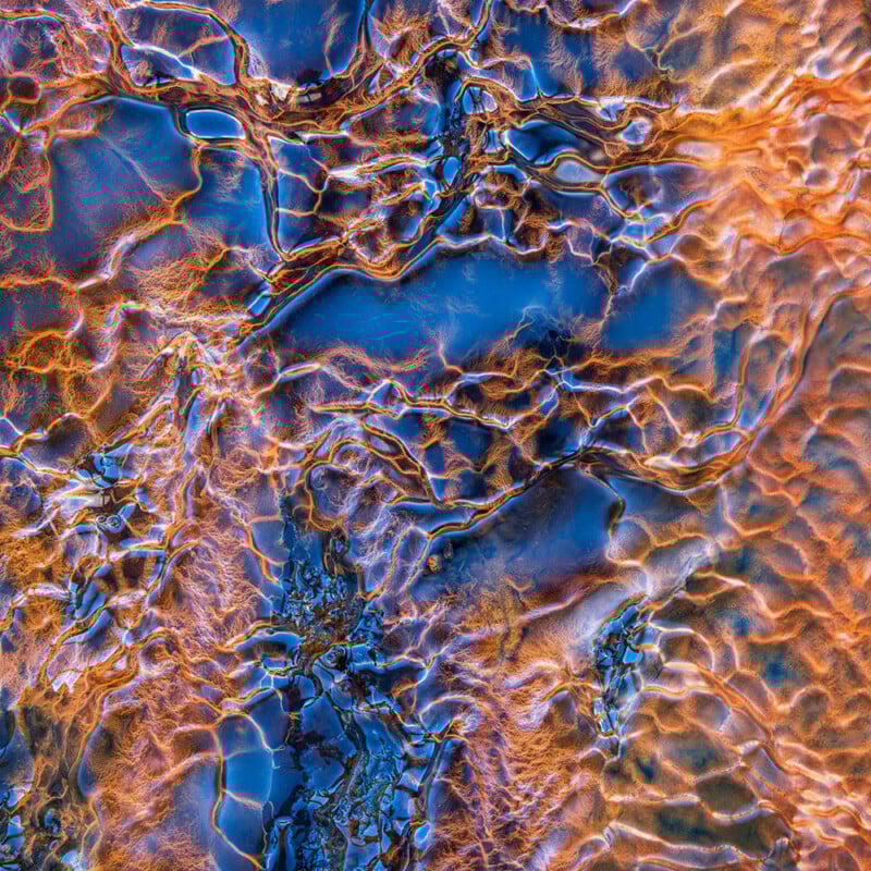 Abstract overhead view of water with rippling patterns reflecting vivid blue and orange light, creating an intricate, web-like texture across the surface.
