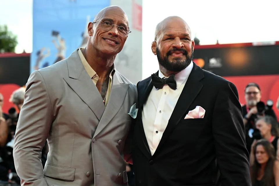 Two men on a red carpet; one in a double-breasted suit, the other in a tuxedo with a bow tie and pocket square