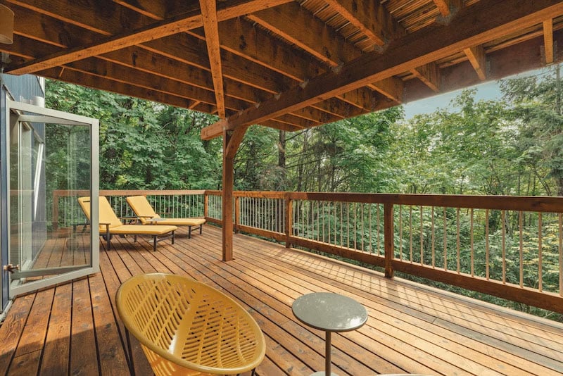 A wooden deck with a slanted roof, surrounded by trees. The deck has two yellow lounge chairs, a small round table, and a yellow woven chair. Glass doors are open to the outdoor space.