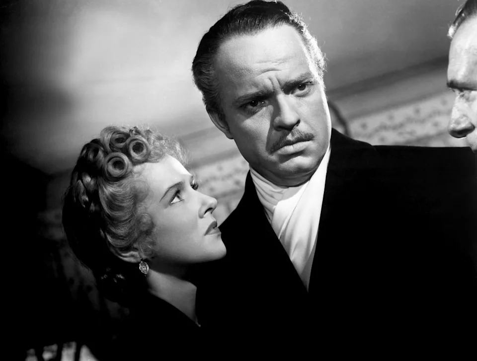 A classic film scene with a woman looking up at a stern man in a suit with a mustache, both in formal attire from an old Hollywood movie