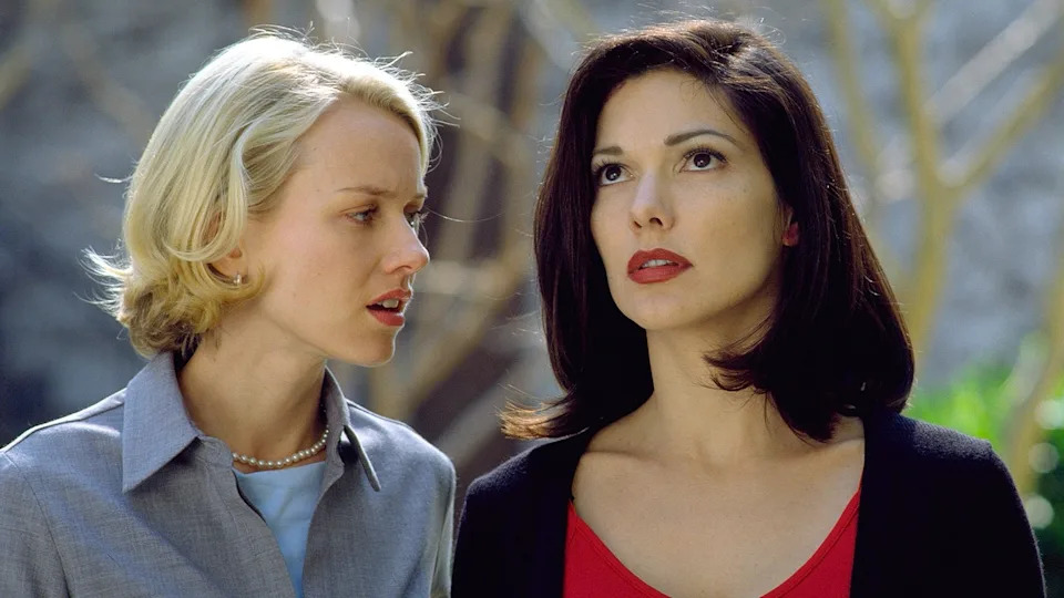 Two women from a movie scene, one with short hair and pearls, the other with shoulder-length dark hair, both appearing serious and focused