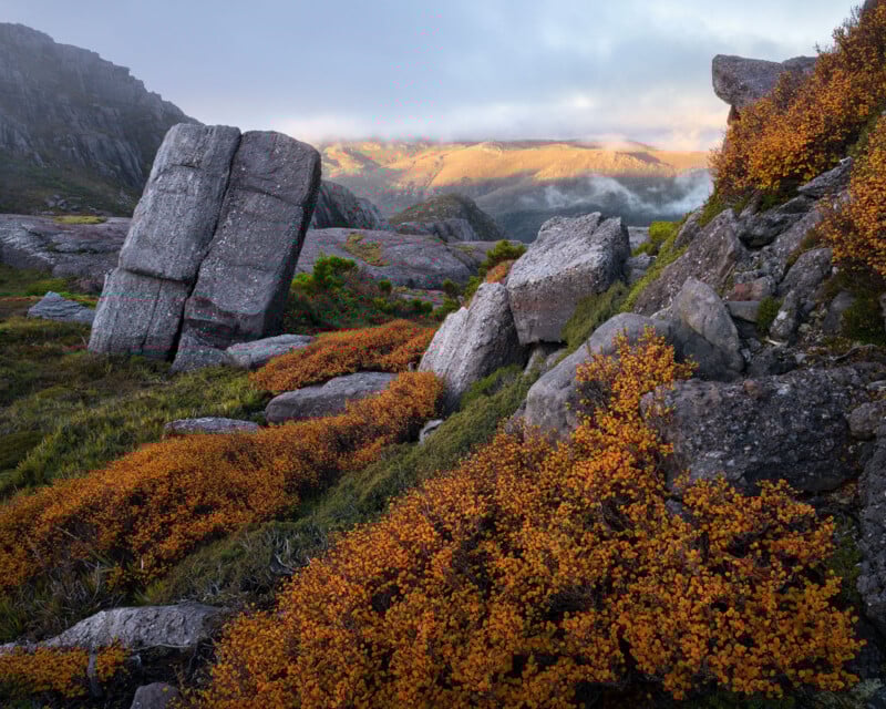 Large rocky boulders and cliffs sit among vibrant orange shrubs on a grassy hillside, with mist and sunlight illuminating distant mountains under a partly cloudy sky.