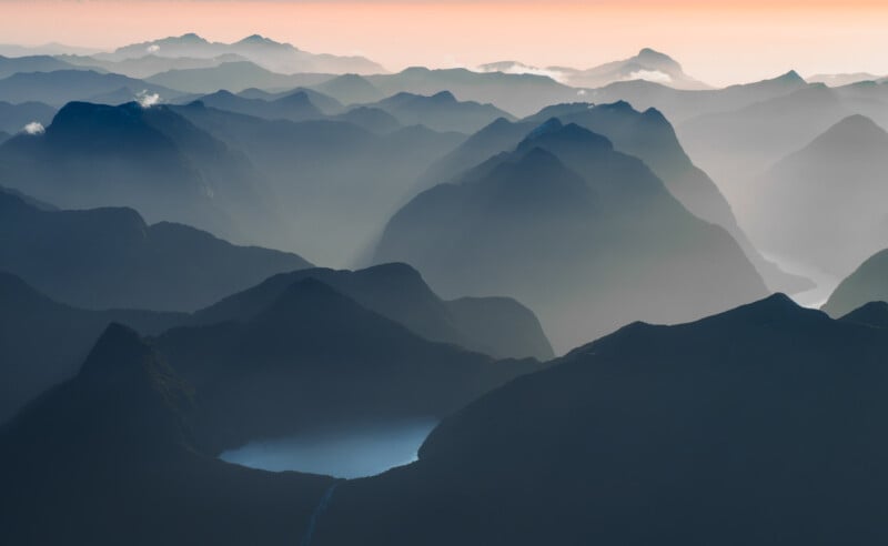 Aerial view of misty mountain ranges at dawn, with layers of dark ridges and soft light illuminating valleys and distant peaks under a pastel orange and blue sky.