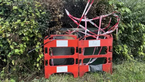 BBC A communications box blackened as the result of a fire, with red safety barriers in front of it and red and white fire tape around it. The box is in front of several green bushes.