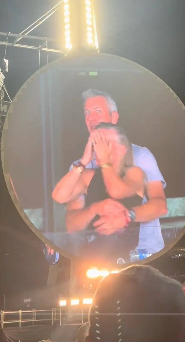 Andy Byron wraps his arms around Kristin Cabot as they appear on a concert screen, with Cabot covering her face in surprise.