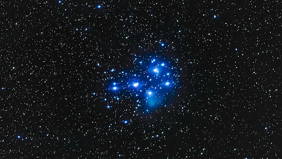 A collection of bright blue glowing stars is pictured in the center of an image hanging against a dense starfield of lower-magnitude stars framed by the blackness of space.