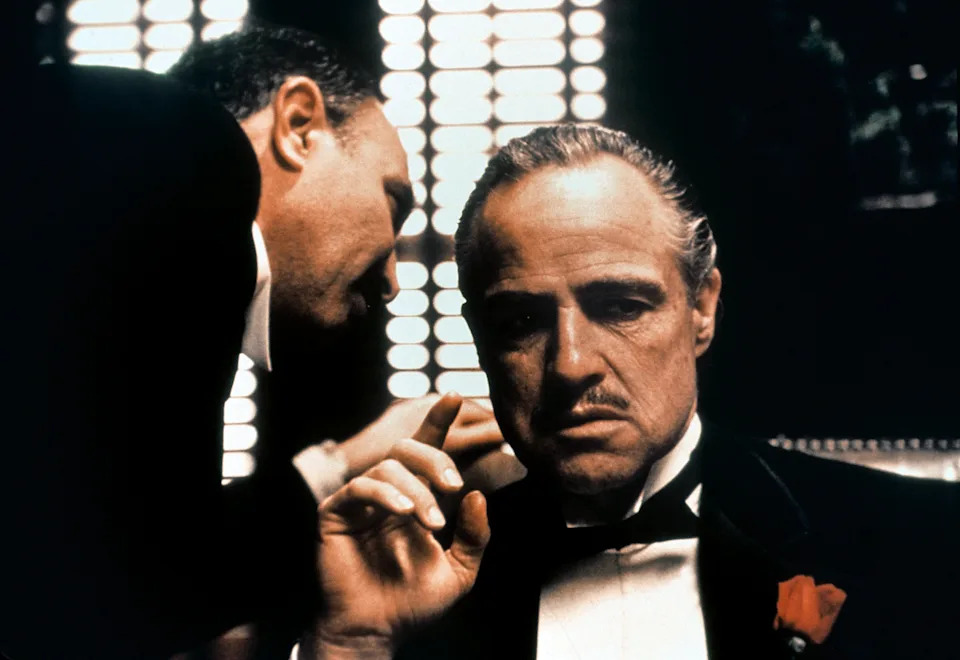 A serious man in a tuxedo listens intently while another whispers in his ear, suggesting a scene of intrigue or power dynamics