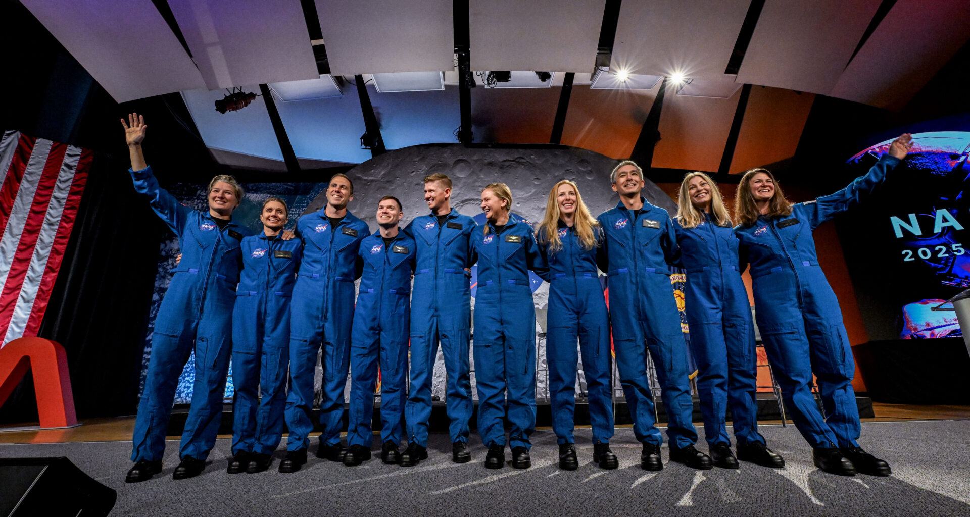 Ten people in blue jumpsuits link arms. The two women on either end wave at the audience, who is not visible.