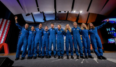 Ten people in blue jumpsuits link arms. The two women on either end wave at the audience, who is not visible.