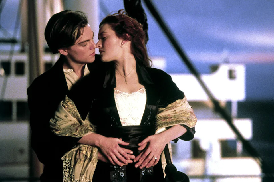 A couple embraces on a ship; the man in a suit and the woman in an elegant dress with a shawl, reminiscent of an iconic movie scene