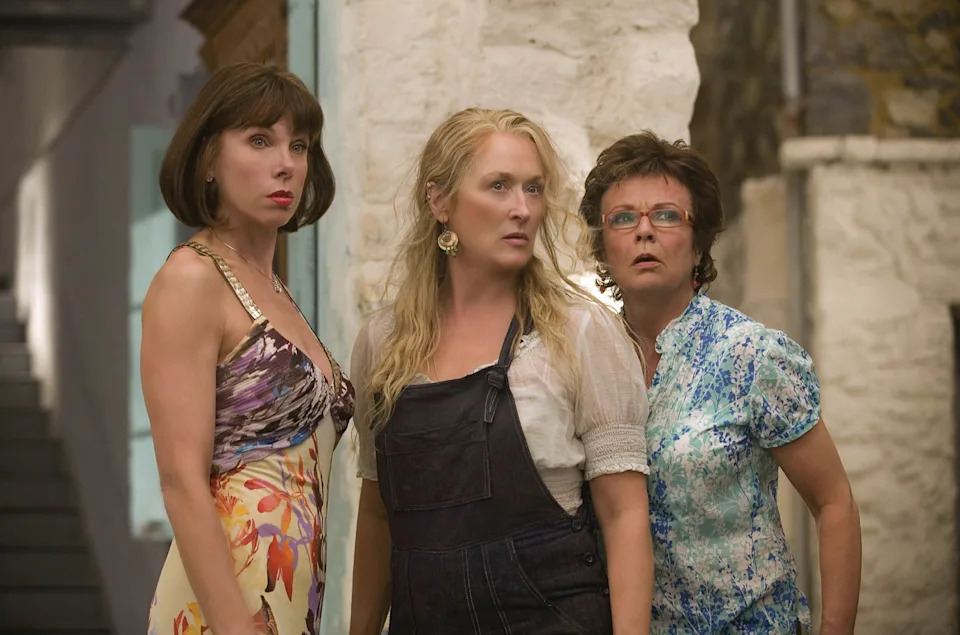 Three women stand together, looking surprised. One wears a floral dress, another wears overalls, and the third wears a patterned blouse