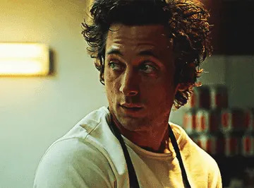 Person with curly hair and an apron looks over their shoulder in a kitchen