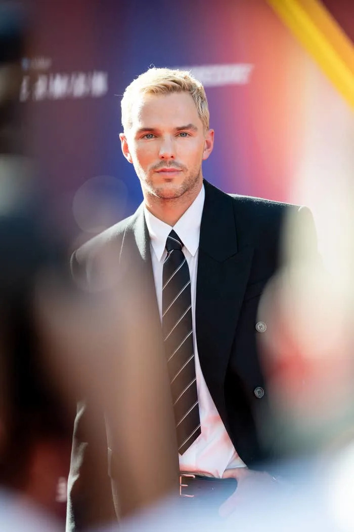 Nicholas Hoult on the red carpet wearing a black suit jacket, white shirt, and striped tie, with short hair styled neatly