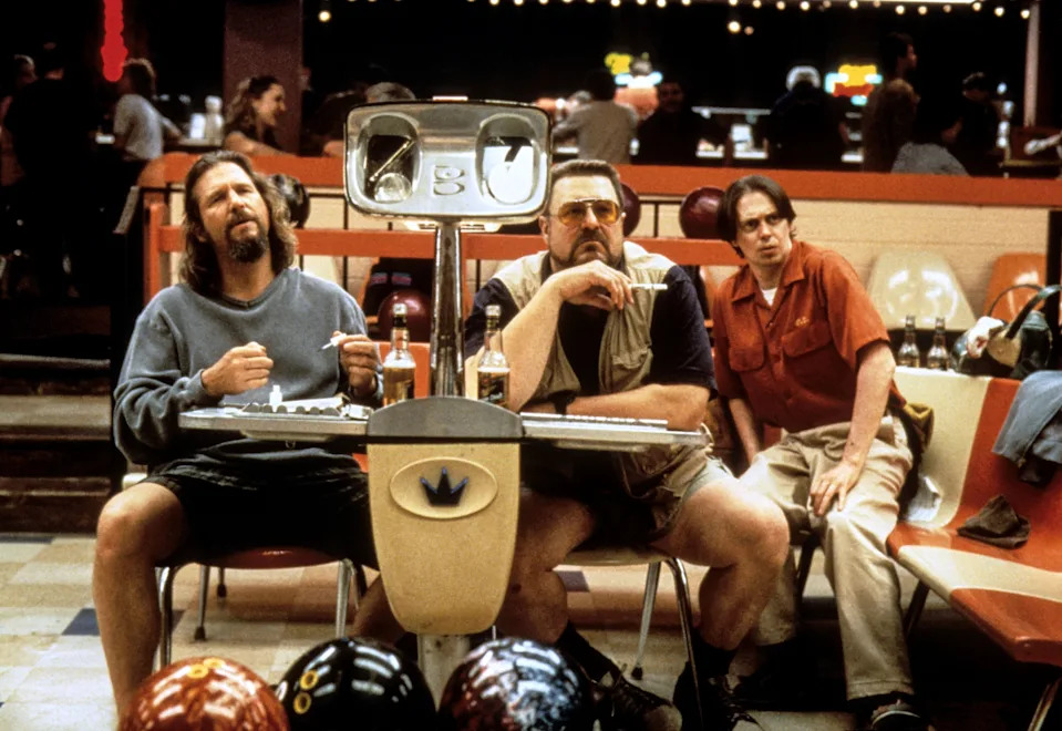 Three men sit in a bowling alley. One wears a robe, another a vest and shorts, and the third a red shirt, all appearing relaxed and focused