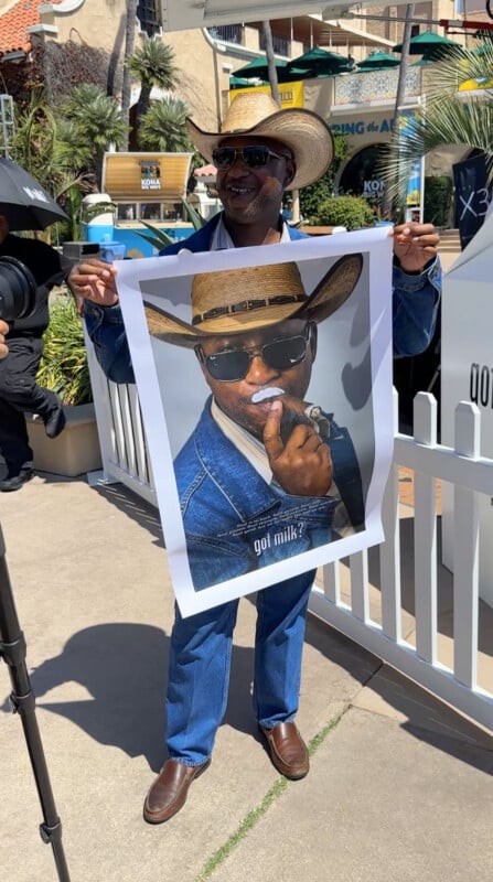 A man in a cowboy hat, sunglasses, and denim clothes smiles while holding a large poster of himself with a milk mustache and the text "got milk?" at an outdoor event.
