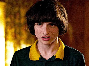 A young person with dark hair, wearing a polo shirt with a yellow collar, looks forward with a surprised expression