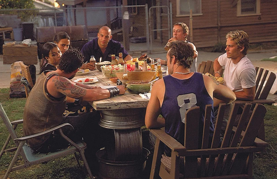 Group of people sit around an outdoor table with food and drinks, engaged in conversation, in a casual setting