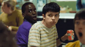 Two teenagers in a cafeteria turn their heads in surprise while holding lunch items