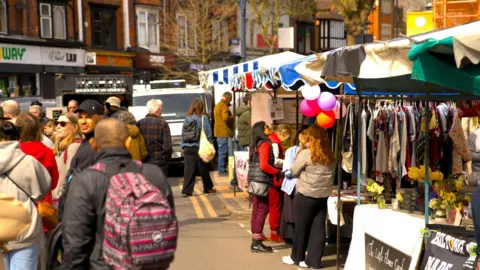 Moseley Farmers' Market A general view of the market with crowds of people walking around the stalls that line both sides of the street. There are shops in the distance too. 