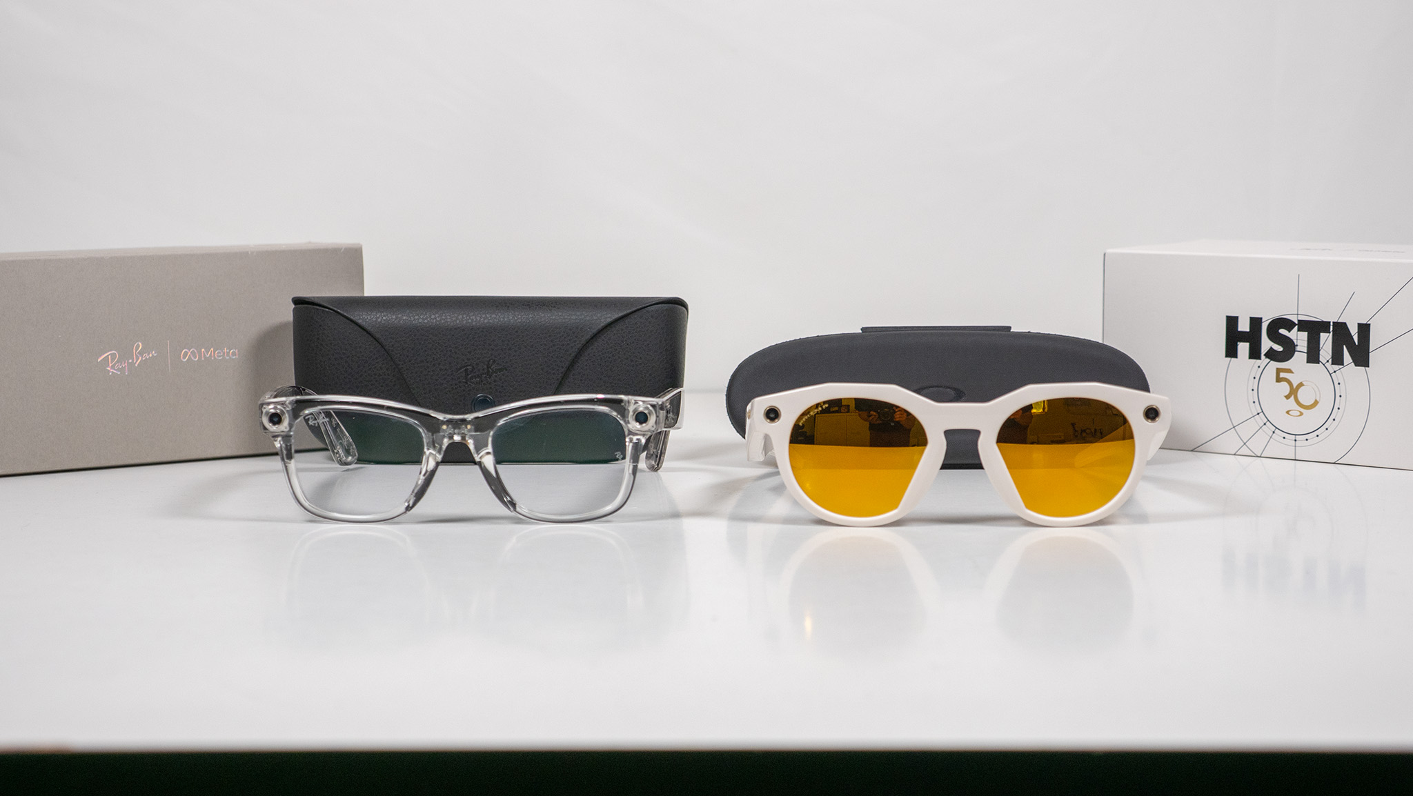Transparent Ray-Ban Meta Smart Glasses and warm white Oakley Meta HSTN smart glasses with their respective boxes and charging cases on a white table