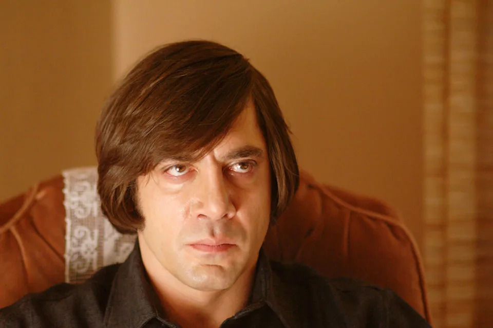 A person with shoulder-length hair and an intense expression, wearing a dark collared shirt, sits in a vintage-style chair