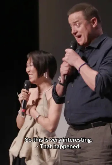 Two people on stage speaking into microphones, one in an elegant sleeveless top, the other in a collared shirt. Subtitles included: "So this is very interesting."