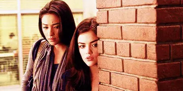 Two women peek around a brick wall with curious expressions. They have long hair and wear casual jackets, appearing to be in a suspenseful situation