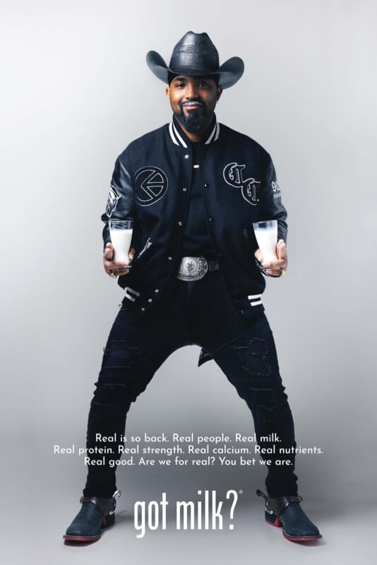 A man in a cowboy hat and black jacket sits on a stool holding two glasses of milk, smiling. Text below reads: “got milk?” Above it, smaller text highlights the benefits of real milk.