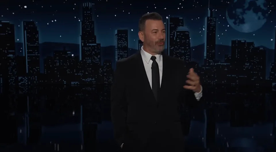 Man in a suit gesturing during a talk show against a cityscape night backdrop