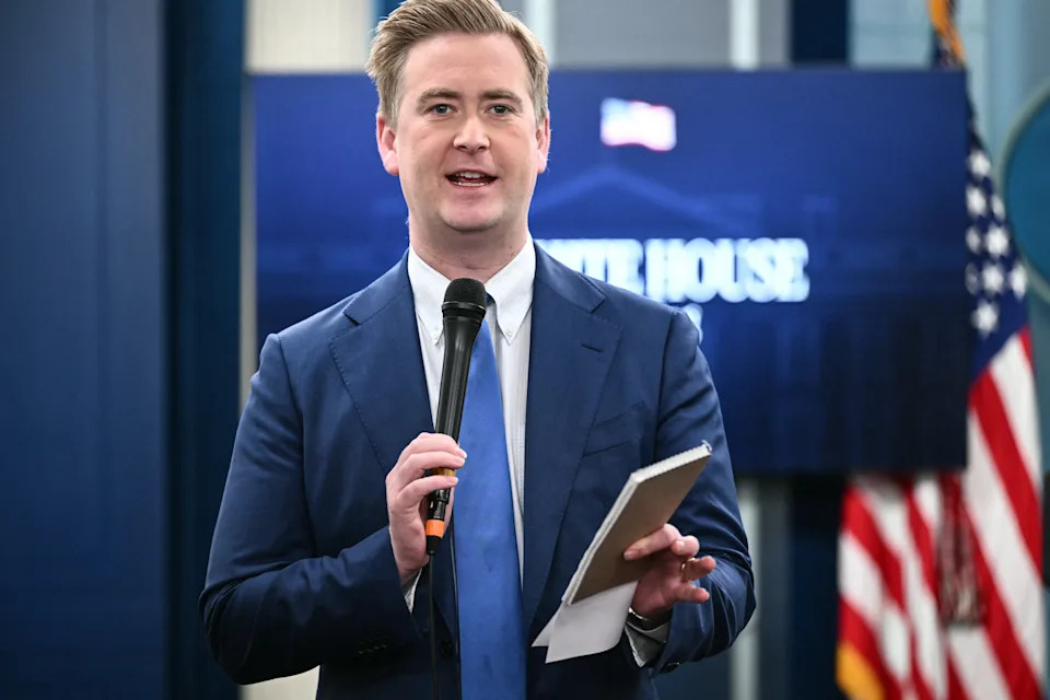 Person in a suit holding a microphone and notepad, standing in front of a blurred background with a sign and US flag visible
