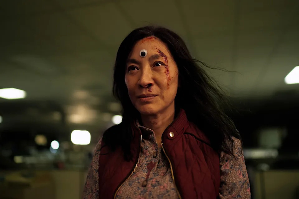 A person with a googly eye on their forehead, wearing a patterned shirt and vest, looks ahead determinedly in a dimly lit setting